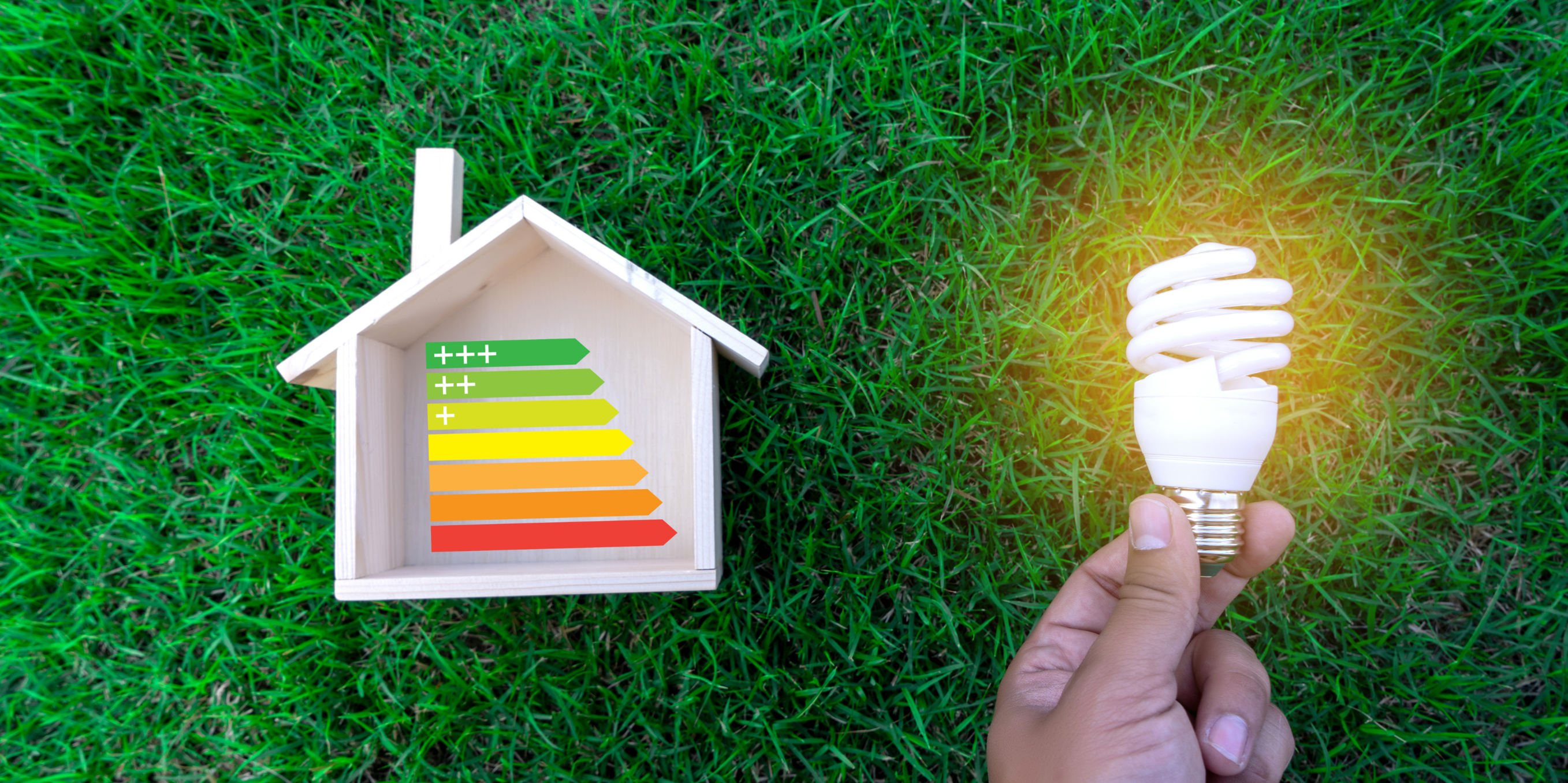 A hand holding an energy-efficient light bulb in front of a house with various energy ratings, symbolizing the importance of choosing energy-efficient appliances to save money and reduce energy consumption. The background is a grassy landscape.